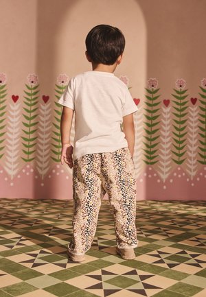 Jeune enfant se tenant face à une fresque florale, portant un t-shirt blanc, un pantalon à motifs et des chaussures beiges sur un sol carrelé géométrique.