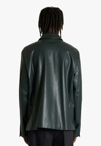 Giacca verde in ecopelle con finitura lucida, colletto con risvolto e vestibilità larga. La vista posteriore mette in evidenza la texture liscia e le cuciture.