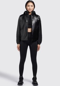 Chaqueta cortavientos negra y brillante con capucha, puños elásticos y cierre delantero; combinada con leggings negros y zapatillas deportivas.