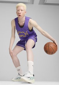 Camiseta de baloncesto de malla morada con gráfico amarillo, pantalones cortos a juego, calcetines blancos y zapatillas negras/blancas. Sosteniendo un balón de baloncesto.