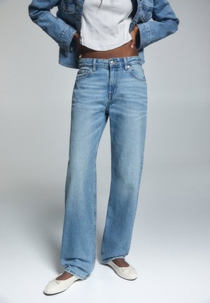 Person iført lyseblå vidde bukser, hvid cropped tank top, denimjakke og hvide ballerinaer, stående mod en ensfarvet baggrund.