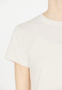 Persona con una camiseta de manga corta de cuello redondo en un tono beige claro, mostrada desde el hombro hasta la mitad del torso, contra un fondo blanco.
