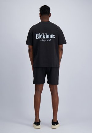 Man staat met de rug naar de camera, draagt een zwart T-shirt met de tekst "Blckbnns Design Dept." en zwarte korte broek tegen een grijze achtergrond.