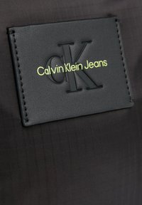 Parcha de tela negra con el logo en relieve "CK" y el texto "Calvin Klein Jeans" en verde brillante. Textura suave con un patrón sutil.