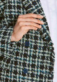 Hand, die den Ärmel einer Tweedjacke mit blauen, weißen und schwarzen Karos und blauen Knöpfen über einem weißen Hemd anpasst.