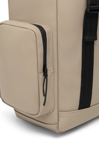 Beige hátizsák oldalsó zsebbel, fekete cipzárral, texturált anyaggal és kontrasztos fekete pánttal. Simább felület és letisztult vonalak.