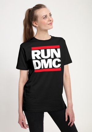 Młoda kobieta z kucykiem, ubrana w czarny t-shirt Run DMC, patrzy w górę i uśmiecha się na jednolitym białym tle.