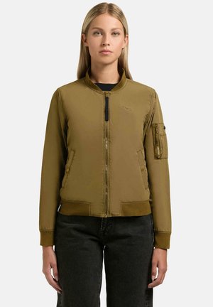 Kobieta ubrana w oliwkowo-zieloną kurtkę bomber z przednim zamkiem błyskawicznym, kieszenią na lewym rękawie, ściągaczami na mankietach i u dołu, w połączeniu z czarnymi spodniami.