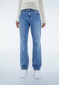 Světle modré džíny s uvolněným střihem, pěti kapsami a viditelným šitím. Spárováno s krémovým pleteným svetrem a bílými botami.