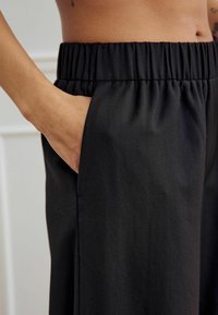 Käsi mustade elastsete vöödega pükste taskus, mille kandjal on nähtav kõht ja käe tätoveering.