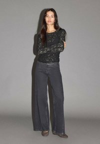 Crna čipkasta majica dugih rukava s okruglim izrezom, u kombinaciji sa svijetlosivim širokim trapericama s metalnim detaljima na pojasu.