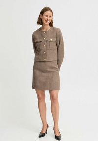 Chaqueta marrón texturizada con cierre de botones y dos bolsillos en el pecho, acompañada de una falda a juego. Diseñado con elegantes tacones altos negros.