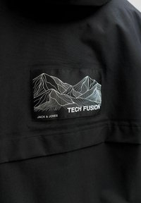 Must mustrik, millel on õmmeldud ristkülikukujuline plekk, kus on näha valged kontuurjooned mägedest ja tekst "Tech Fusion" ning "Jack & Jones."