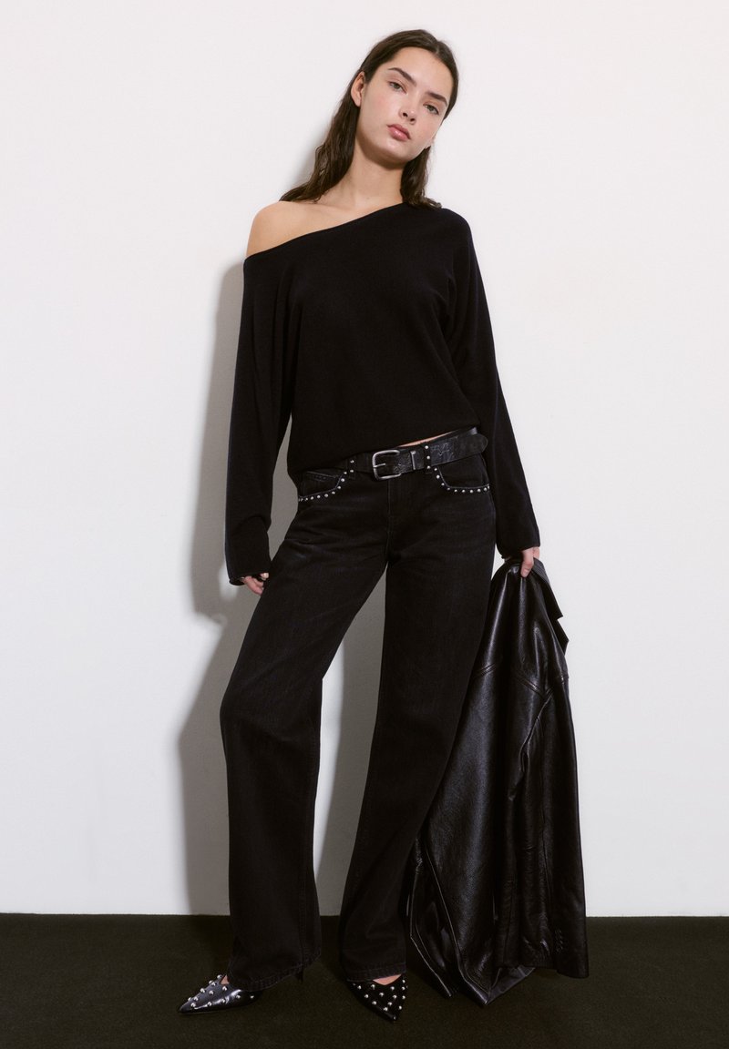 Schwarzer oversized Pullover mit einem freigelegten Schulter, kombiniert mit hochgeschnittenen schwarzen Jeans mit Nietenakzenten und spitzen schwarzen Schuhen.