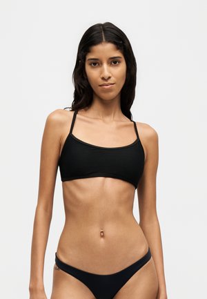 Jeune femme aux longs cheveux foncés portant un haut de bikini côtelé noir et un bas de bikini noir assorti sur un fond clair uni.