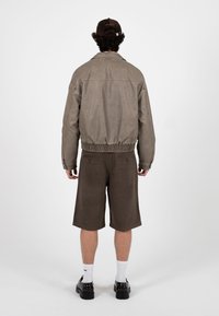 Mand iført brun kasket, taupe jakke med elastisk taljebånd, mørkebrune knælange shorts, hvide sokker og sorte sko, stående med ryggen til.