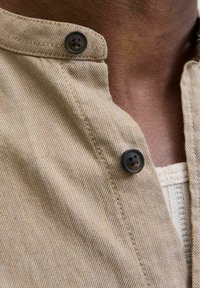 Tan striped shirt collar with two black buttons, showing soft fabric texture and subtle stitching details. Features a layered look with an inner shirt.
