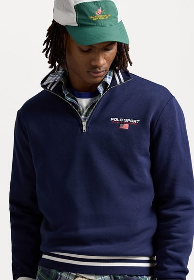 Námořnický modrý pulovr s čtvrtím zipem a bílým a modrým pruhovaným límcem. Vyšitý logo "Polo Sport" a detail americké vlajky na hrudi.