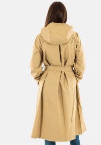 Lange beige regenjas met capuchon, een ceintuur in de taille en een rugplooi. Gemaakt van glad, waterproof materiaal voor buitengebruik.