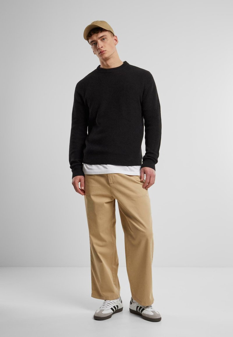 Black ribbed sweater over a white t-shirt, paired with beige wide-leg pants and white sneakers with black stripes; beige cap on head.
