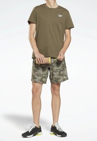 Osoba v olivovém tričku Reebok, maskáčových šortkách a černých teniskách, držící hnědou a žlutou láhev na vodu se šňůrou.