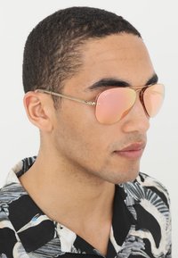 Óculos de sol aviador com armação dourada e lentes espelhadas em rosa, exibindo um design elegante e superfície refletiva. Camisa floral casual ao fundo.