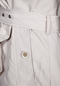 Lichtbeige trenchcoat met een gestructureerd ontwerp, stoffen riem, grote voorkantzak met een witte knop en gedetailleerde stiksels.