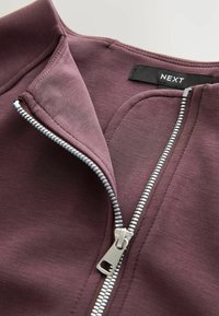 Tumši sarkans apģērbs ar rāvējslēdzēju, gluda tekstūra, sudraba krāsas rāvējslēdzējs un birka ar uzrakstu "NEXT" pie kakla.