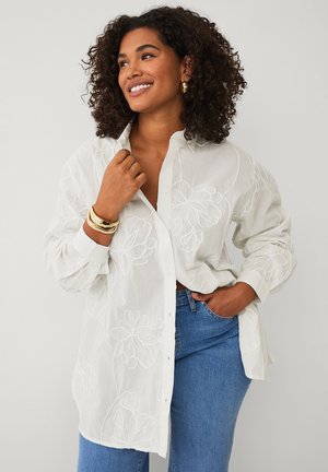 Smilende kvinde med krøllet hår iført hvid skjorte med blomsterbroderi, blå jeans, guldøreringe og armbånd, poserer mod en ensfarvet baggrund.