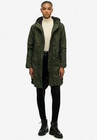 Parka vert olive avec une capuche, fermeture éclair avant et cordon de serrage à la taille. Associée à des leggings noirs et des bottes noires à lacets. Texture lisse.