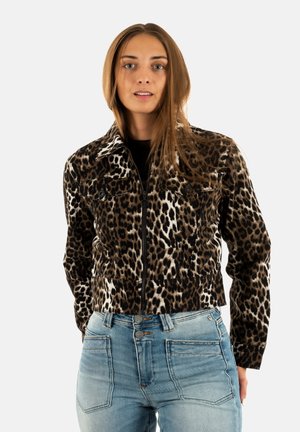 Giovane donna con i capelli lunghi che indossa una giacca con zip a stampa leopardata e jeans a vita alta di un blu chiaro, su uno sfondo semplice.