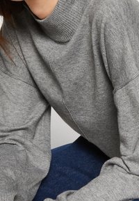 Pull en tricot gris avec un col côtelé haut et des épaules tombantes, présentant une texture douce et une coupe décontractée.