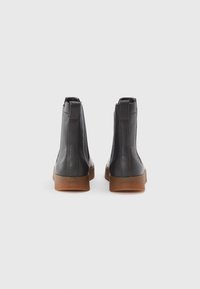 Bottines en cuir noir avec une finition texturée, semelles en caoutchouc marron contrastantes et zips latéraux. Présente un branding subtil au niveau du talon.