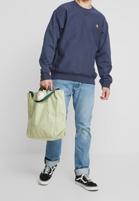 Man i blå sweatshirt och jeans, hållande en ljusgrön totebag, iförd svarta och vita sneakers, stående framför en enfärgad bakgrund.
