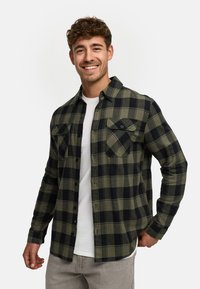 Chemise en flanelle à carreaux verts et noirs avec un devant boutonné, deux poches poitrine et des manches longues, portée par-dessus un T-shirt blanc.