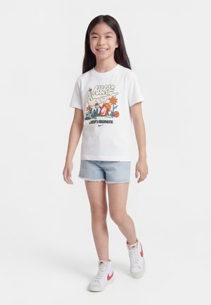 Glimlachend meisje met lang donker haar, gekleed in een wit Nike T-shirt met opdruk, lichte denim shorts en witte Nike hoge sneakers, lopend naar voren.