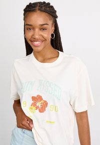 T-shirt couleur crème avec un graphique floral coloré et du texte. Manches courtes, coupe décontractée et matériau en coton doux.