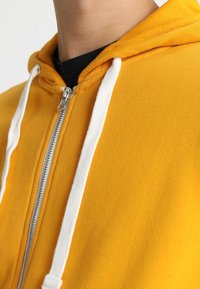 Sweat à capuche jaune zippé avec une finition texturée, doté d'un cordon blanc et d'une fermeture éclair argentée, porté par-dessus une chemise noire.