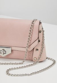 Světle růžová kabelka z umělé kůže s texturovaným povrchem, stříbrným řetízkem a dekorativní kovovou sponou s ostrými designovými prvky.