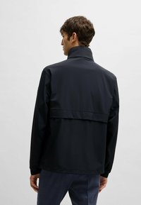 Marinblå vattentät jacka med hög krage, långa ärmar och ett overlay bak; slät textur och minimalistisk design.