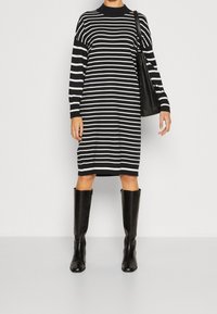 Laivansiniset ja valkoiset raidalliset neulemekot, joissa on korkea kaula-aukko, pitkät hihat ja sivuviillot, yhdistettynä korkeisiin mustiin nahkabootseihin ja mustaan laukkuun.