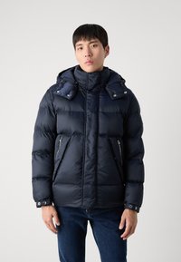 Marinblå pufferjacka med hög krage och avtagbar huva, med dragkedjefickor och ett texturerat mönster. Ankellånga ärmar med knapppryttningar.