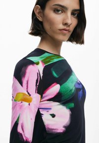 Haut à manches longues noir, orné d'un motif floral éclatant dans les tons de rose, vert et bleu. Le tissu a une texture lisse et une coupe légèrement ajustée.