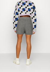 Person som bär svart-vita rutiga shorts, en färgglad rutiga kortärmad tröja och vita sneakers, stående mot en enfärgad bakgrund.