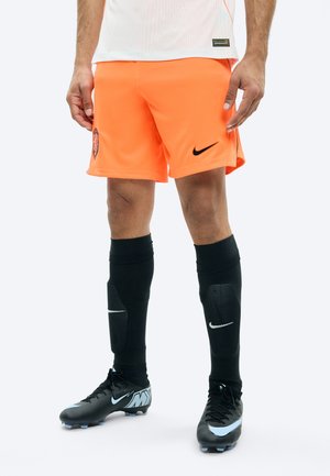 Männliche Beine, die knallorange Nike-Fußballshorts, schwarze Nike Schienbeinschoner und schwarze Nike Fußballschuhe tragen, stehen vor weißem Hintergrund.