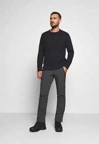 Moški stoji z eno roko v žepu, oblečen v črno pulover, temno sive hlače in črne škornje na enostavni svetli podlagi.