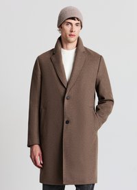 Cappotto di lana marrone con collo a revers, chiusura con un bottone e tasche laterali. Indossato sopra un maglione color crema e abbinato a un berretto beige lavorato a maglia.