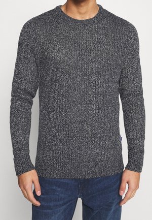 Mężczyzna ubrany w czarno-biały melanżowy sweter z długimi rękawami i ciemnoniebieskie dżinsy, stojący z ramionami swobodnie opuszczonymi wzdłuż ciała.