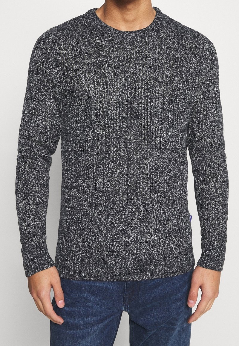 Homme portant un pull en maille noir et blanc à motifs tachetés, à manches longues, et un jean bleu foncé, debout avec les bras détendus le long du corps.