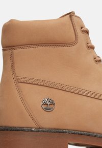 Kožená bota barvy světle hnědé s kulatou špičkou, stříbrným logem, kontrastním prošíváním a dekorativním leskem na okraji podrážky.